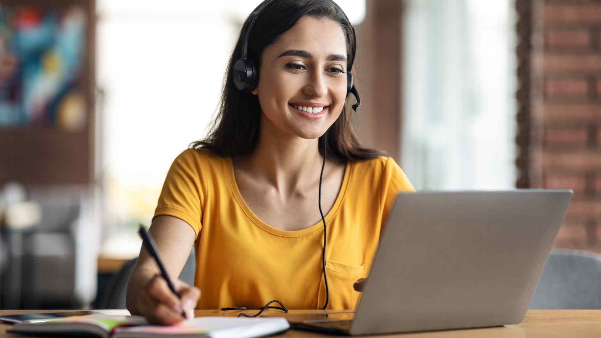 Sarı tişörtlü, kulaklık takan ve bilgisayarı üzerinden online ders alan kadın